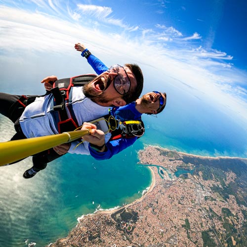 Vue saisissante d'un tandem en chute libre, dos aux Sables d'Olonne, expressions de joie ou de peur sur les visages, sous un ciel d'un bleu intense d'été, avec l'océan Atlantique turquoise en arrière-plan