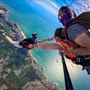 Vue aérienne spectaculaire d'un saut en parachute tandem avec un vidéoman en chute libre face à eux, tenant les mains du moniteur au-dessus de la côte vendéenne, la passagère sourit à la caméra